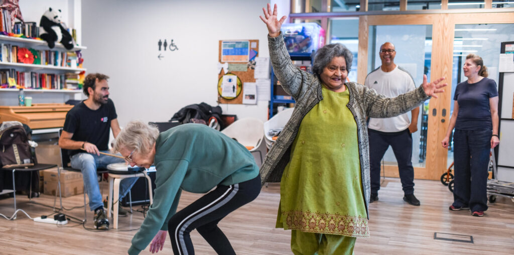 Two older women stretching and dancing in a hall with one man playing the drums.